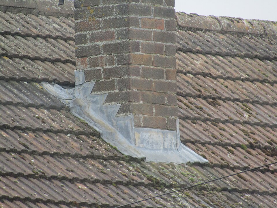 Lead flashing where a chimney stack passes through a tiled roof: an apron below, stepped flashing dressed into the brick joints up each side, so rain runs past the junction.