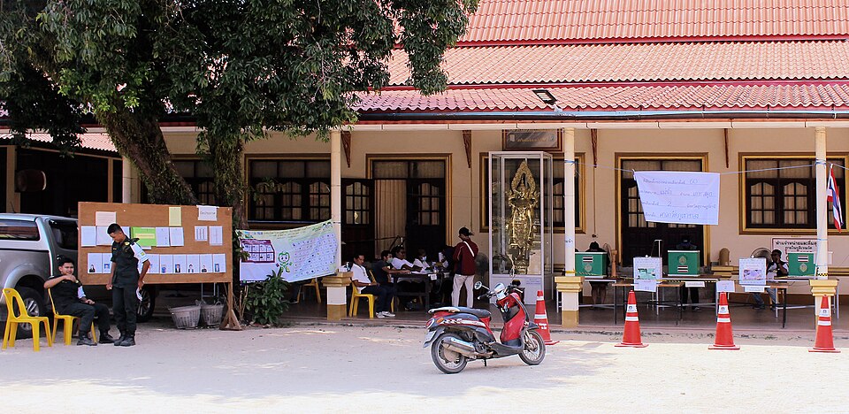 A polling station in Thailand, 2023, laid out in the open: voter lists posted on the board at left, the check-in table, screened booths, and sealed ballot boxes in plain view.