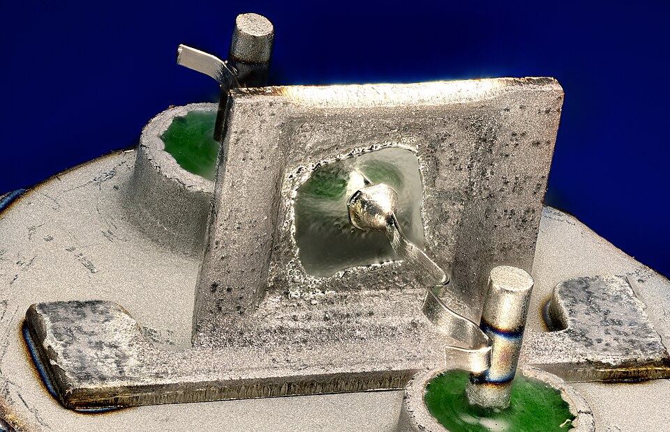 Inside a 2N1307, a 1960s germanium alloy-junction transistor, under magnification: the germanium chip sits in the window of its metal frame, with a fine wire bonded from the metal dot alloyed into its face to the post at the right. Hand-made transistors are this scale and kind.