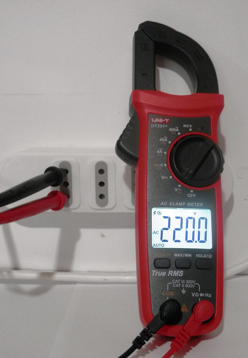 A clamp meter reading 220 V AC across a socket with its test probes. Its jaws measure current without cutting any wire, by closing round a single conductor.
