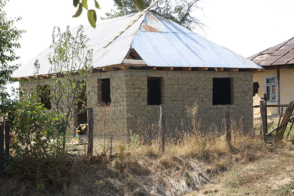A single-storey adobe house in Romania at roof stage, before plastering. Sun-dried blocks laid in running bond, small openings with timber lintels, and a timber wall plate along the wall tops carrying the roof.