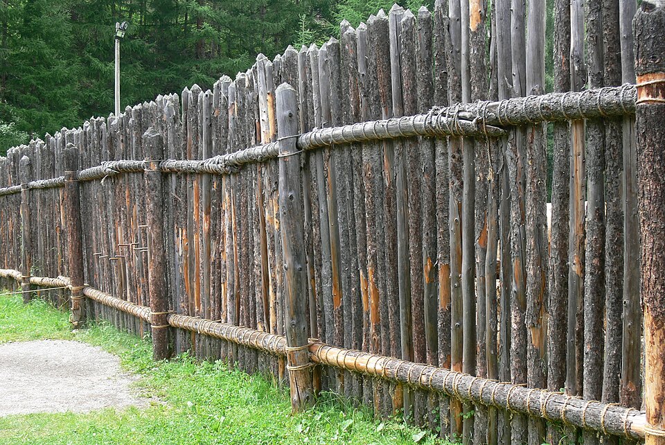 A reconstructed palisade at Archeoparc, Schnalstal: pointed poles set upright in a trench and lashed to horizontal rails. It does not stop a determined attacker; it slows one and forces the approach to the gate.