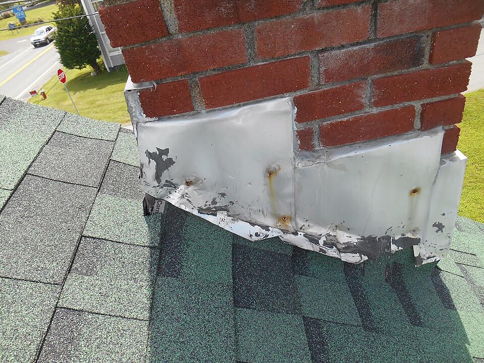 A failed chimney flashing: sheet metal patched on, lifting and rusting, with no step into the brick joints. Water runs behind it into the roof and down the flue.