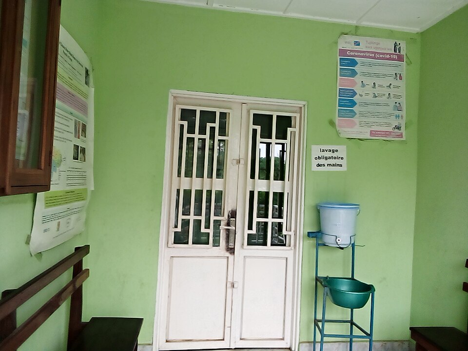 A handwashing station at a building entrance (2023): a bucket with a tap above a basin on a stand, under a sign that makes handwashing compulsory.