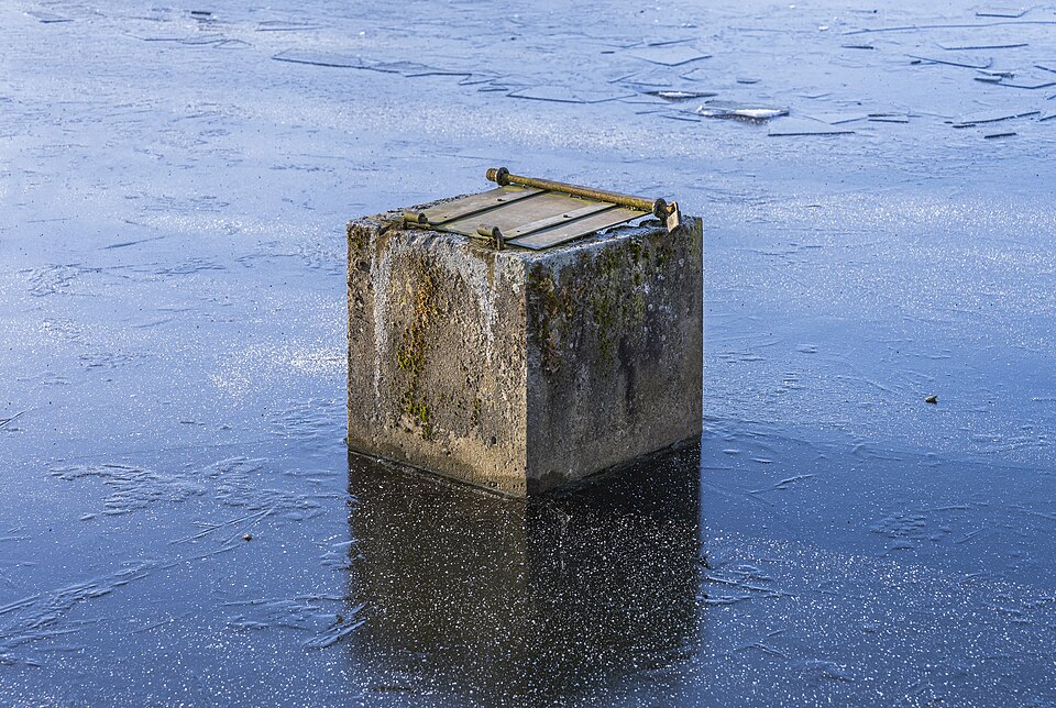 A concrete monk outlet standing in the frozen Knollenteich, Hof, Germany (2024): the square tower stands in the pond, with a cover of boards on top.