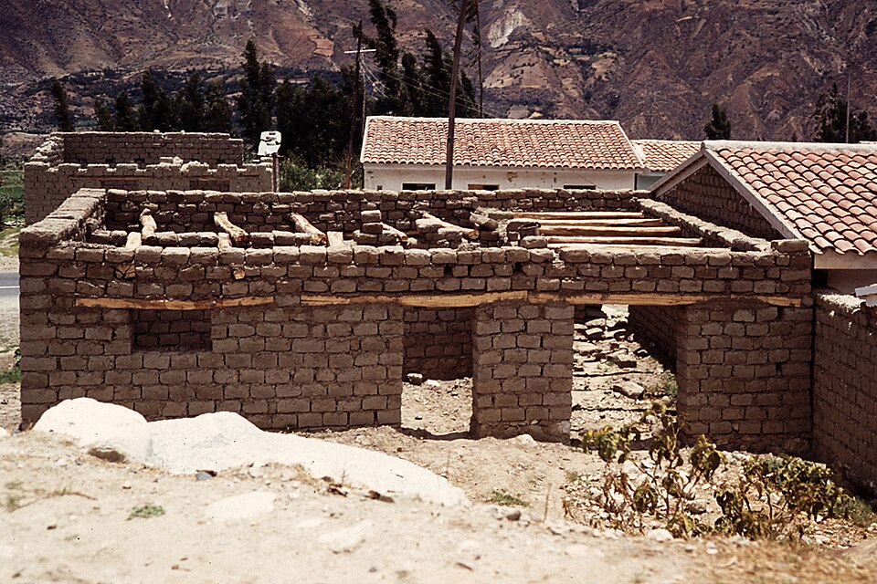 An adobe house under construction in the Peruvian Andes. A continuous timber band runs across the wall tops at lintel height, tying every wall together and spanning the openings.