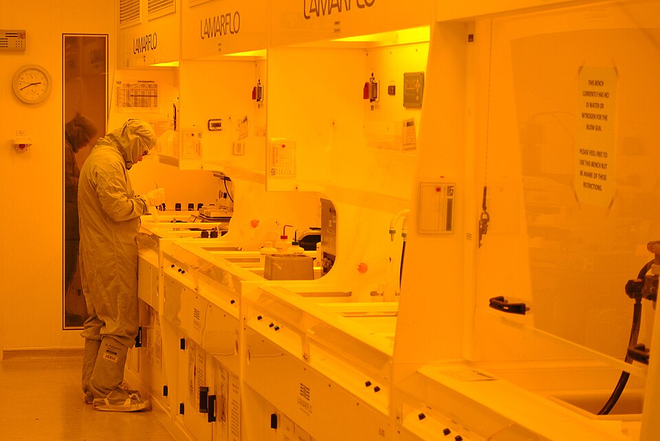 A university photolithography clean room (UCL, 2013). The light is yellow because blue and ultraviolet light would expose the photoresist; the worker is covered head to foot because people shed particles.