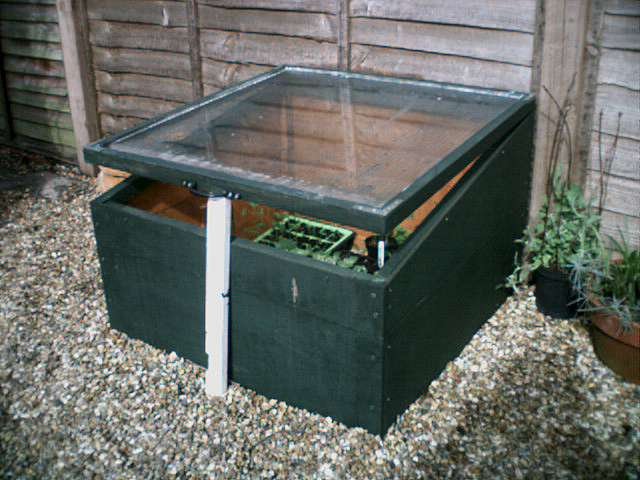 A cold frame: a boarded box with a glazed lid, propped open on a stick to let heat out on a sunny day.