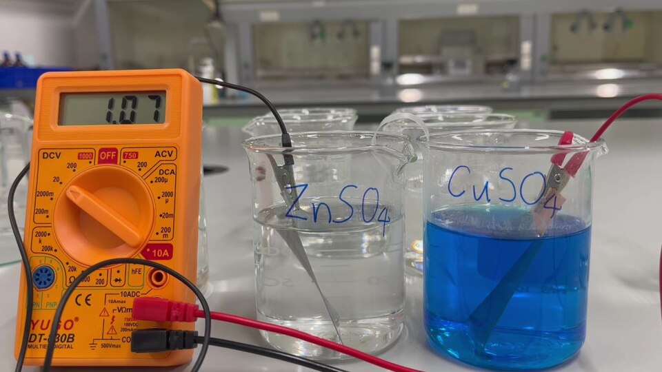 A Daniell cell in a school laboratory: zinc in zinc sulphate, copper in copper sulphate, the two solutions joined by a salt bridge. The meter reads 1.07 volts — close to the 1.1 volts a Daniell cell gives.