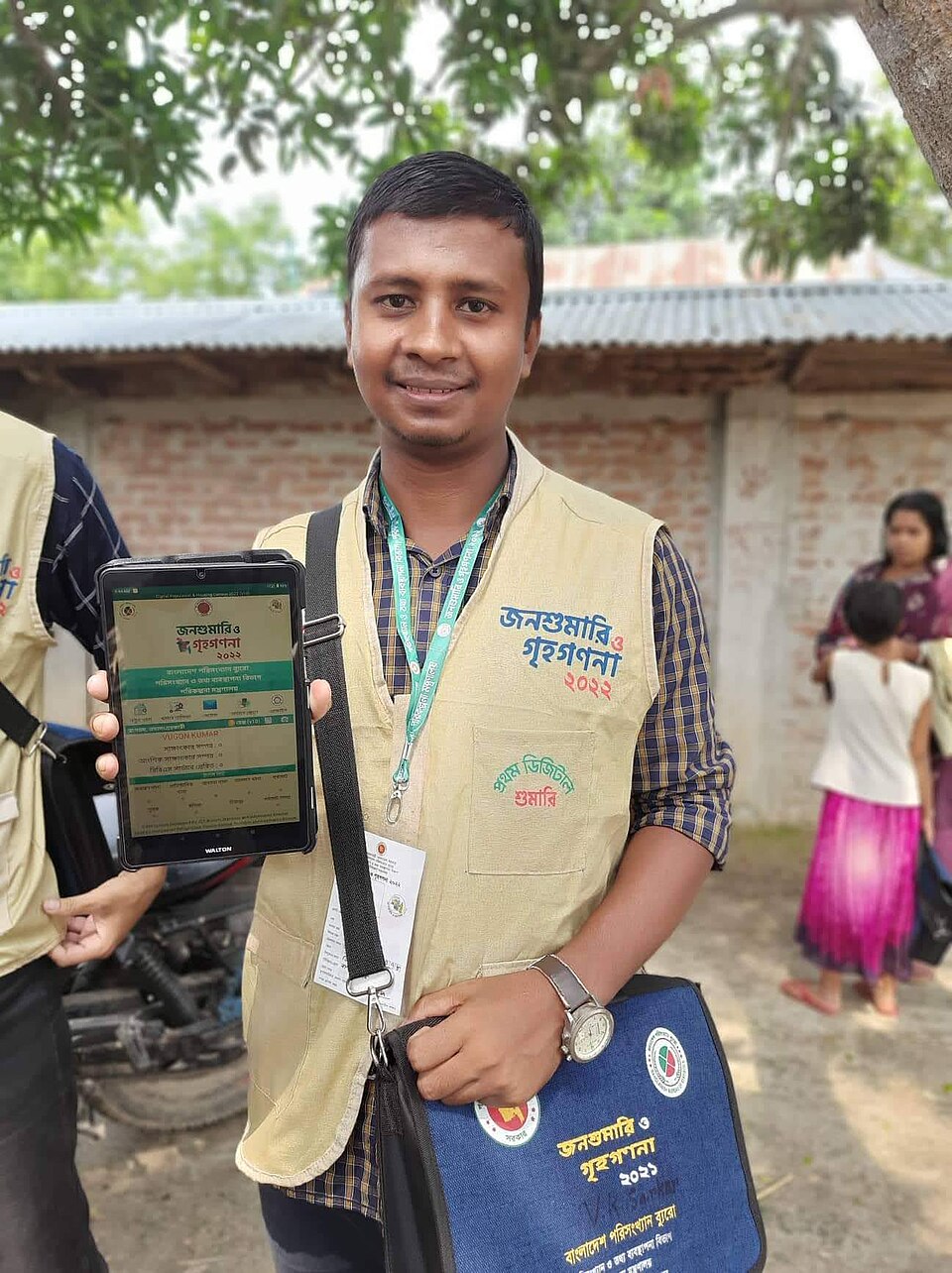A census enumerator in Bangladesh, 2022: identity card on a lanyard, a vest that marks the role, and the questionnaire on a tablet. A paper form does the same job; the visible identity is what gets the door opened.