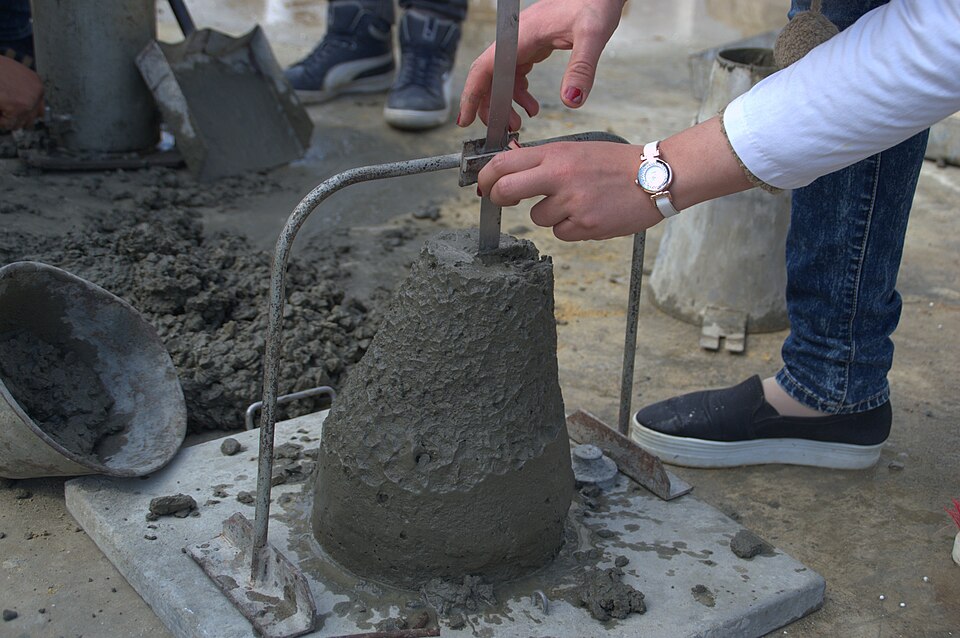 Measuring a slump test (2016): the emptied cone stands upside down beside the concrete, and the drop from its top is read with a rule laid across it.