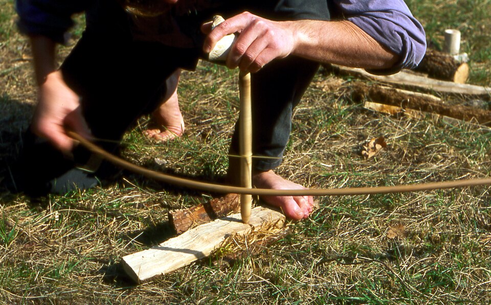 A bow drill in use: bearing block in the left hand, wrist locked to the shin, foot on the board, spindle outside the bow.
