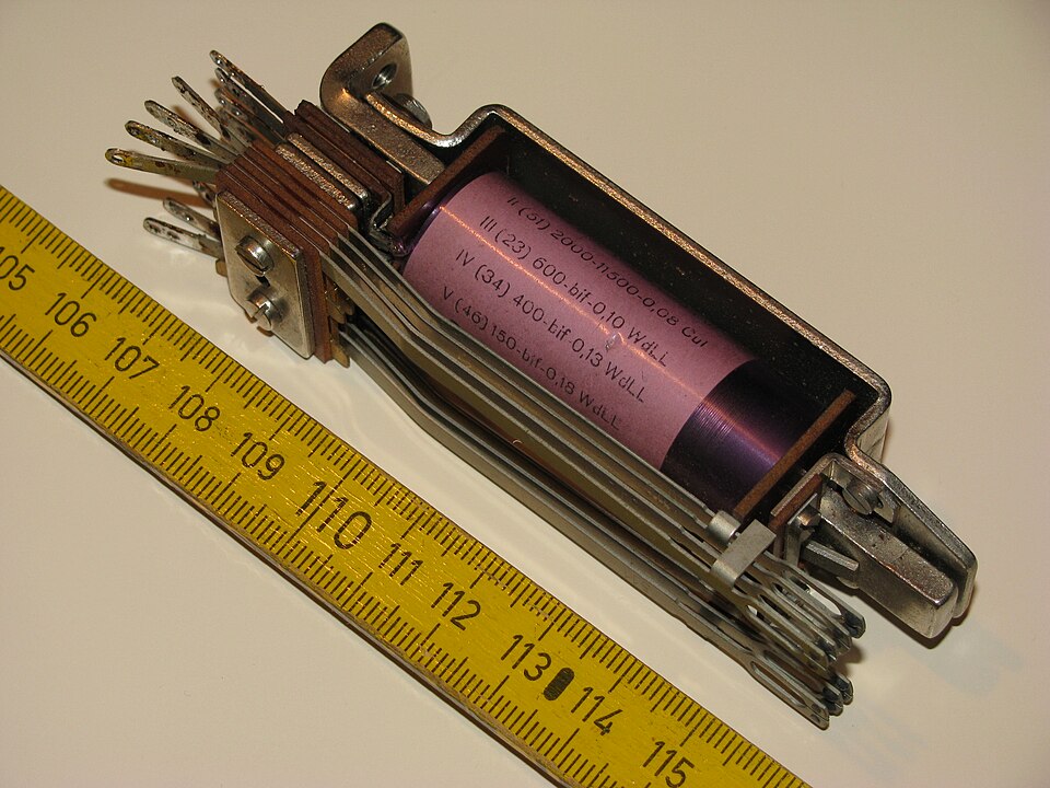 A telephone-exchange relay, about 9 cm long against the rule. Current in the coil (pink label) pulls the armature at the right-hand end, which moves the stack of flat contact springs at the left and opens or closes several circuits at once.