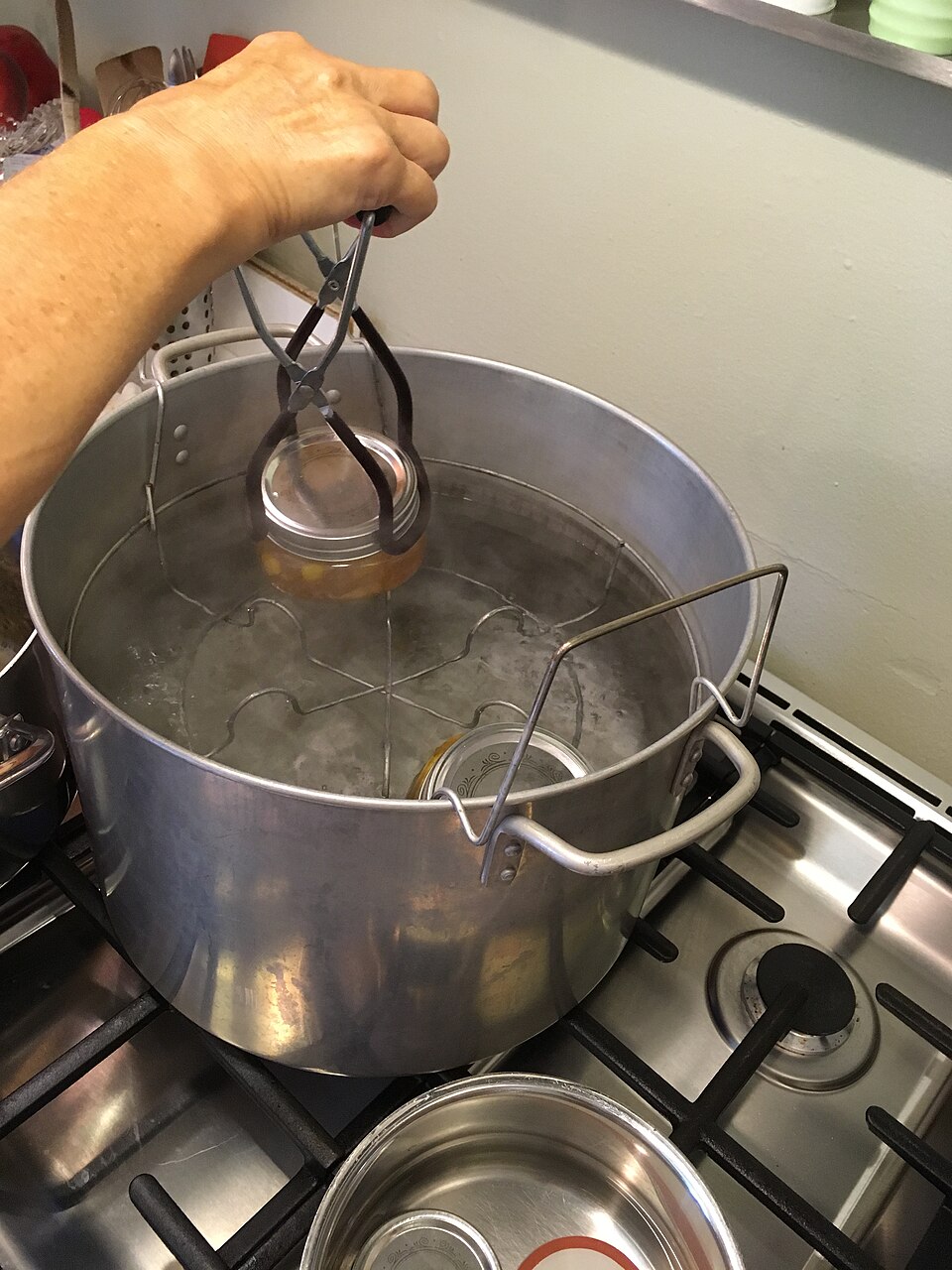 Lowering filled jars into a boiling-water bath with a jar lifter. Safe for acid foods — fruit, jam, vinegar pickles — and never for low-acid vegetables, meat or fish.