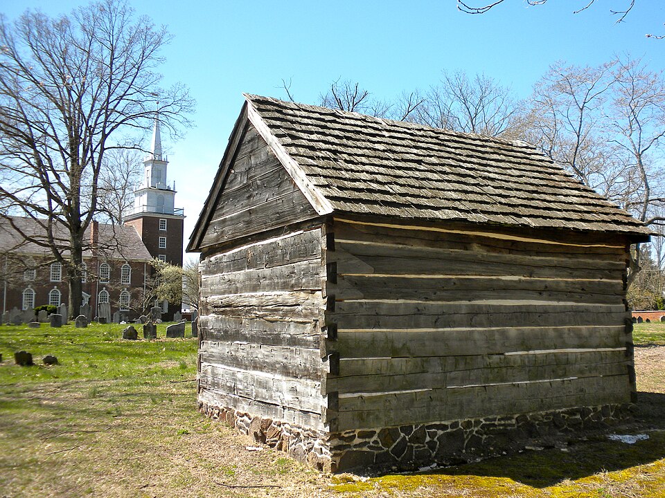 A hewn-log cabin on a stone foundation, with a shake roof and projecting eaves. The stones lift the bottom logs clear of the ground and the splash.