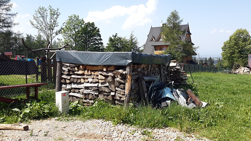 Split firewood stacked under a roof with open sides — air through, rain off. The way firewood actually seasons.