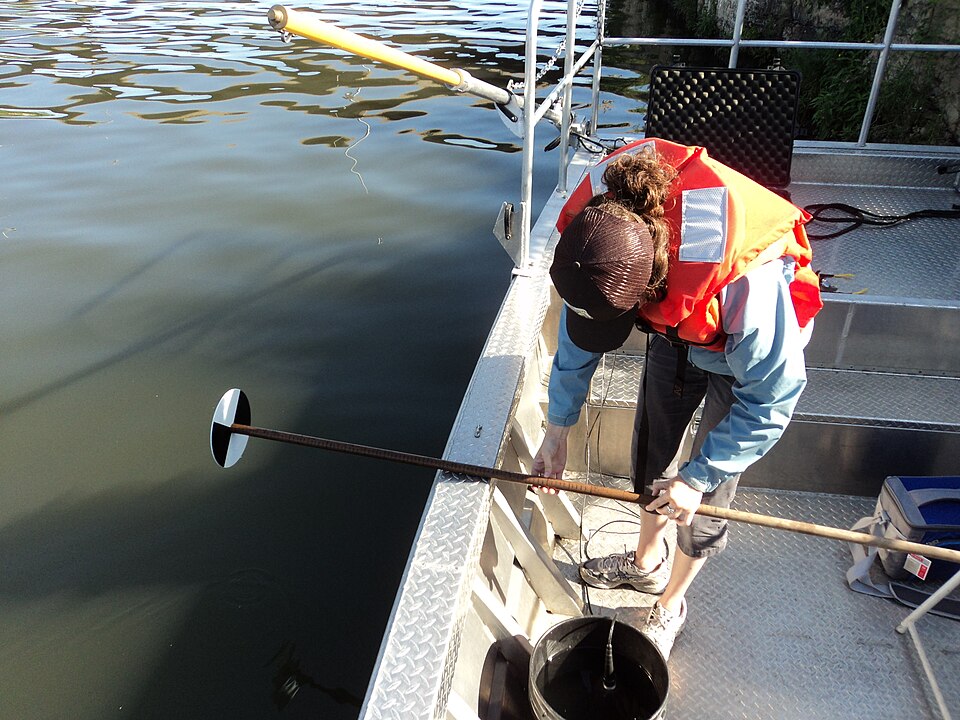 A US Fish and Wildlife Service worker holds a black-and-white Secchi disk on a pole over the water to measure its clarity (2011).
