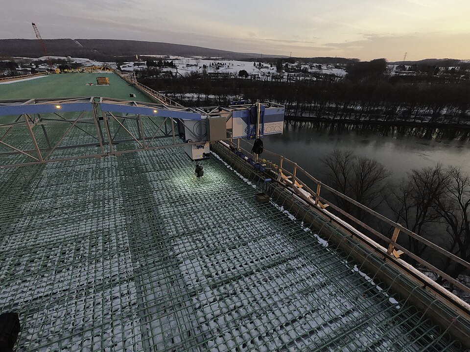 Reinforcing bars laid in mats across a bridge deck before the pour, CSVT River Bridge, Pennsylvania (2020); a gantry robot ties the crossings, and every bar is set on chairs to hold its cover.