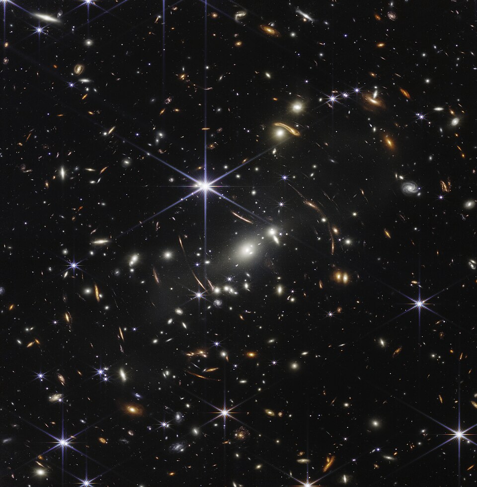 The James Webb Space Telescope's first deep field, 2022: nearly every point of light is a galaxy. The arcs are galaxies far behind the cluster at the centre, their light bent and magnified by its gravity.