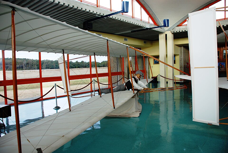 A replica of the Wright 1902 glider in a museum: biplane wings, the elevator in front, the rudder behind, the pilot lying on the lower wing.