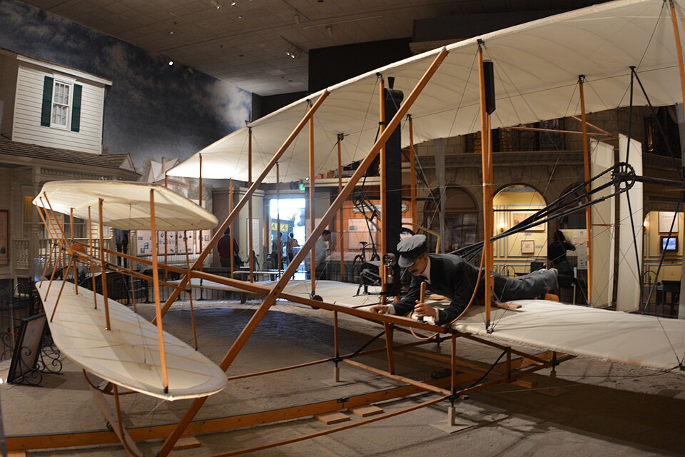 A replica of the 1903 Wright Flyer: the pilot prone on the lower wing beside the engine, chains to two pusher propellers, the elevator in front.