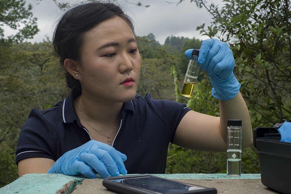 A public-health specialist checks the chlorine level of a village water source in La Paz, Honduras, 2019: the reacted sample vial is held up to the light, with an unreacted blank vial standing beside it for comparison.