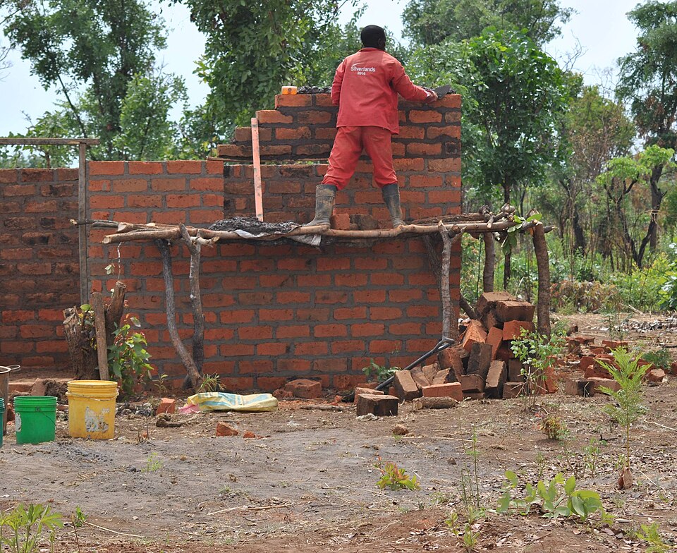 A bricklayer in Zambia raising a house wall in fired brick from a pole scaffold, 2017. Each course is laid to a string line and the joints are broken by half a brick.